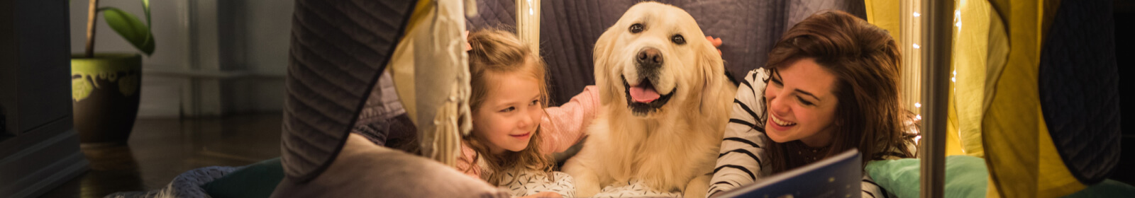 Mother, daughter, and dog laying in a fort smiling while reading a book