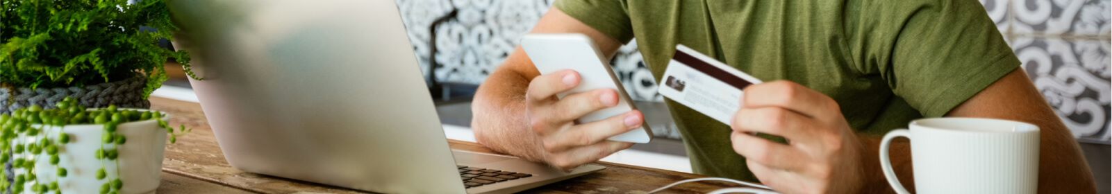 Man holding smart phone and debit card while seated at his table with his laptop and coffee